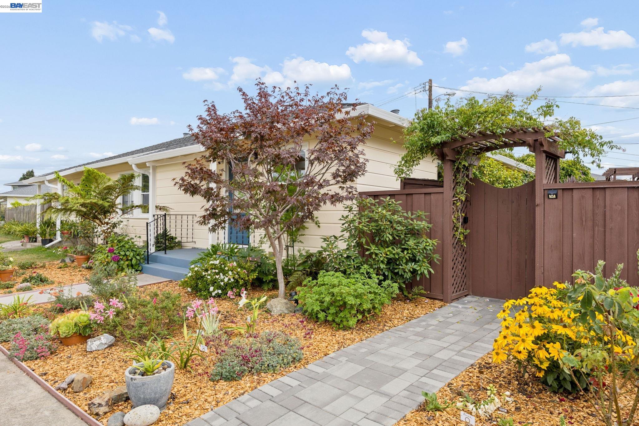 Single-story home with a blue entry porch, flowering garden, paver walkway, and vine-covered wooden arbor.