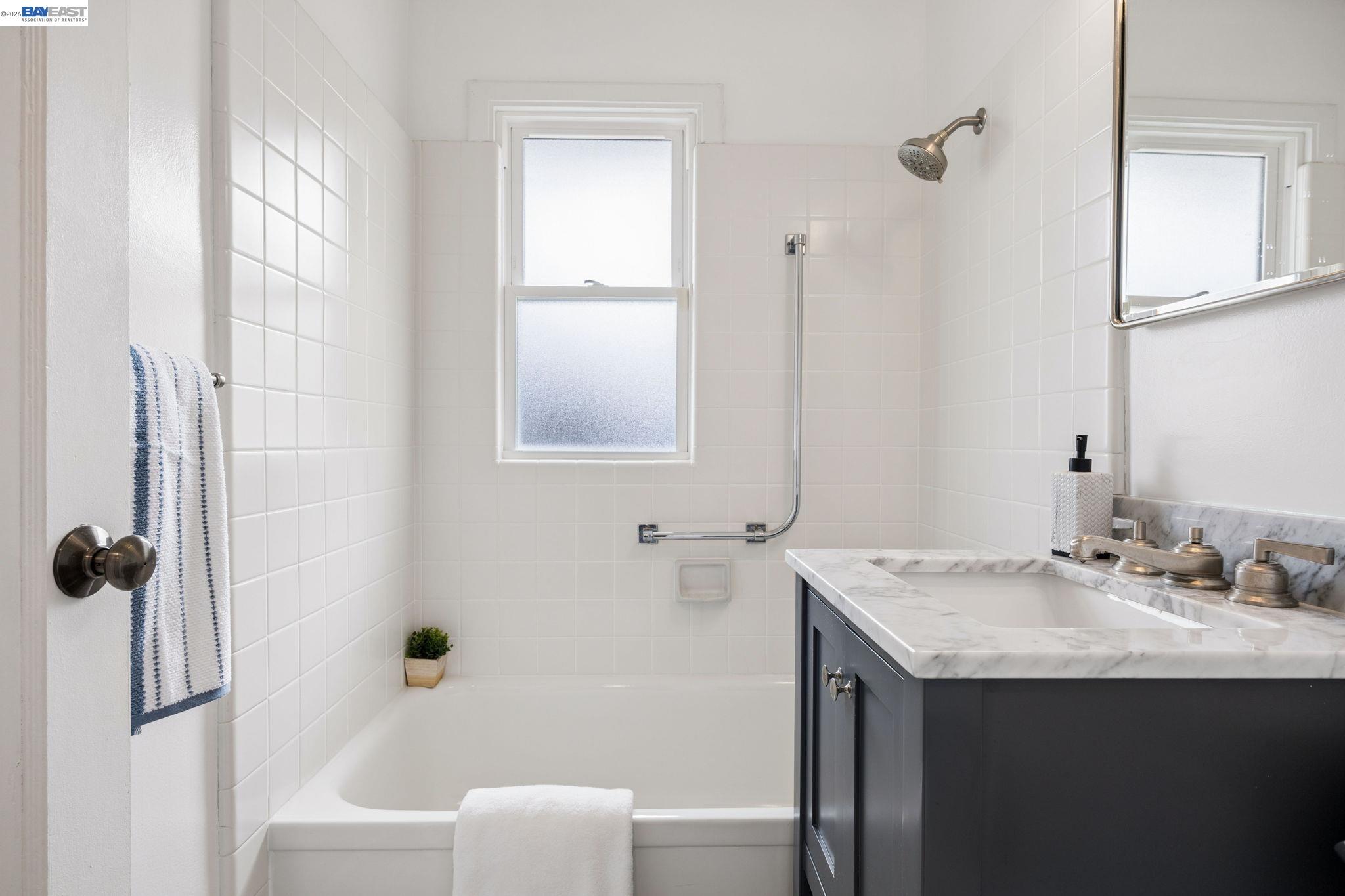 Bathroom with a white tiled tub and shower beside a dark vanity with a marble countertop.