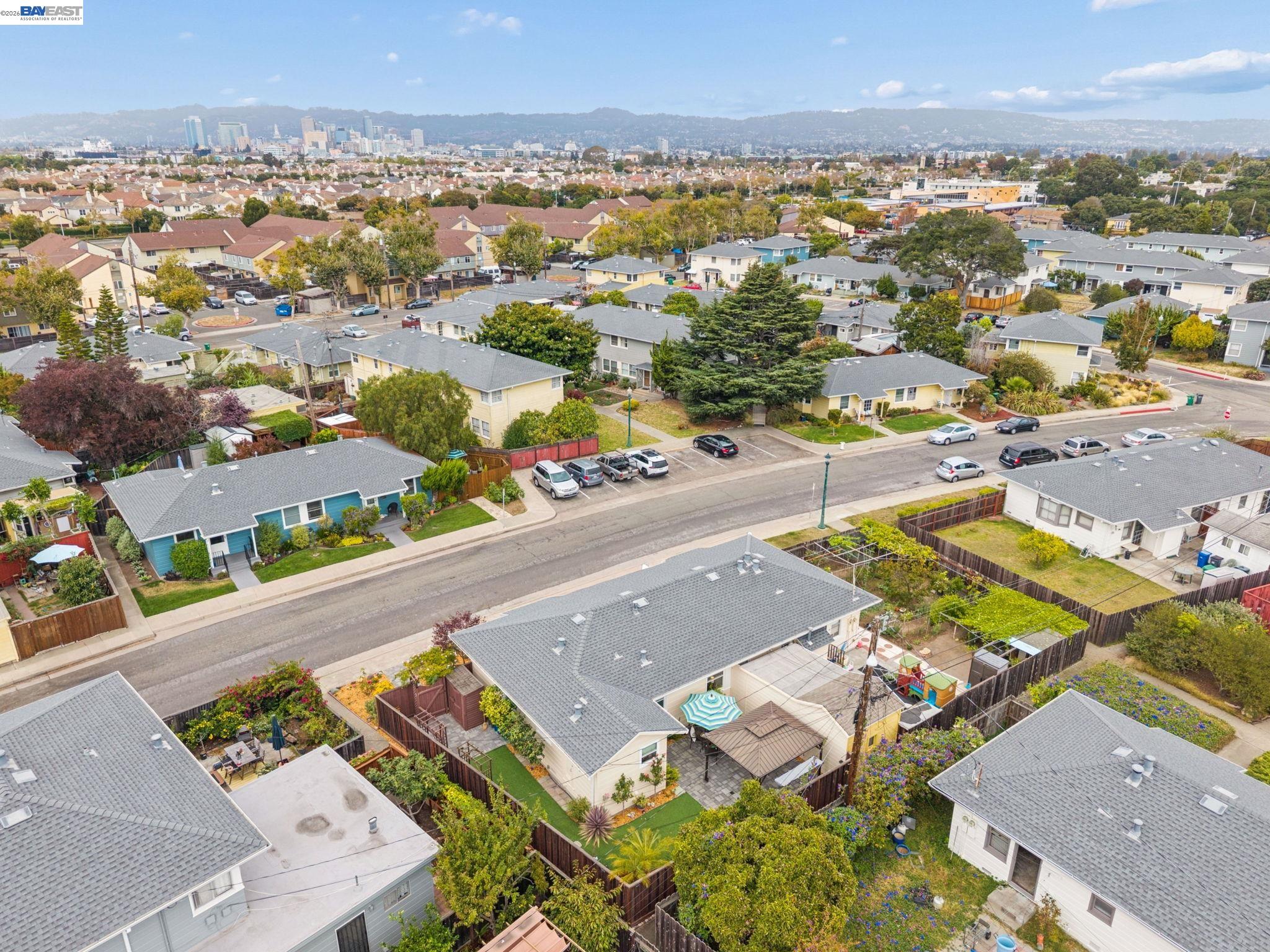 Aerial view of homes and backyards along a residential street, with a distant city skyline and hills.