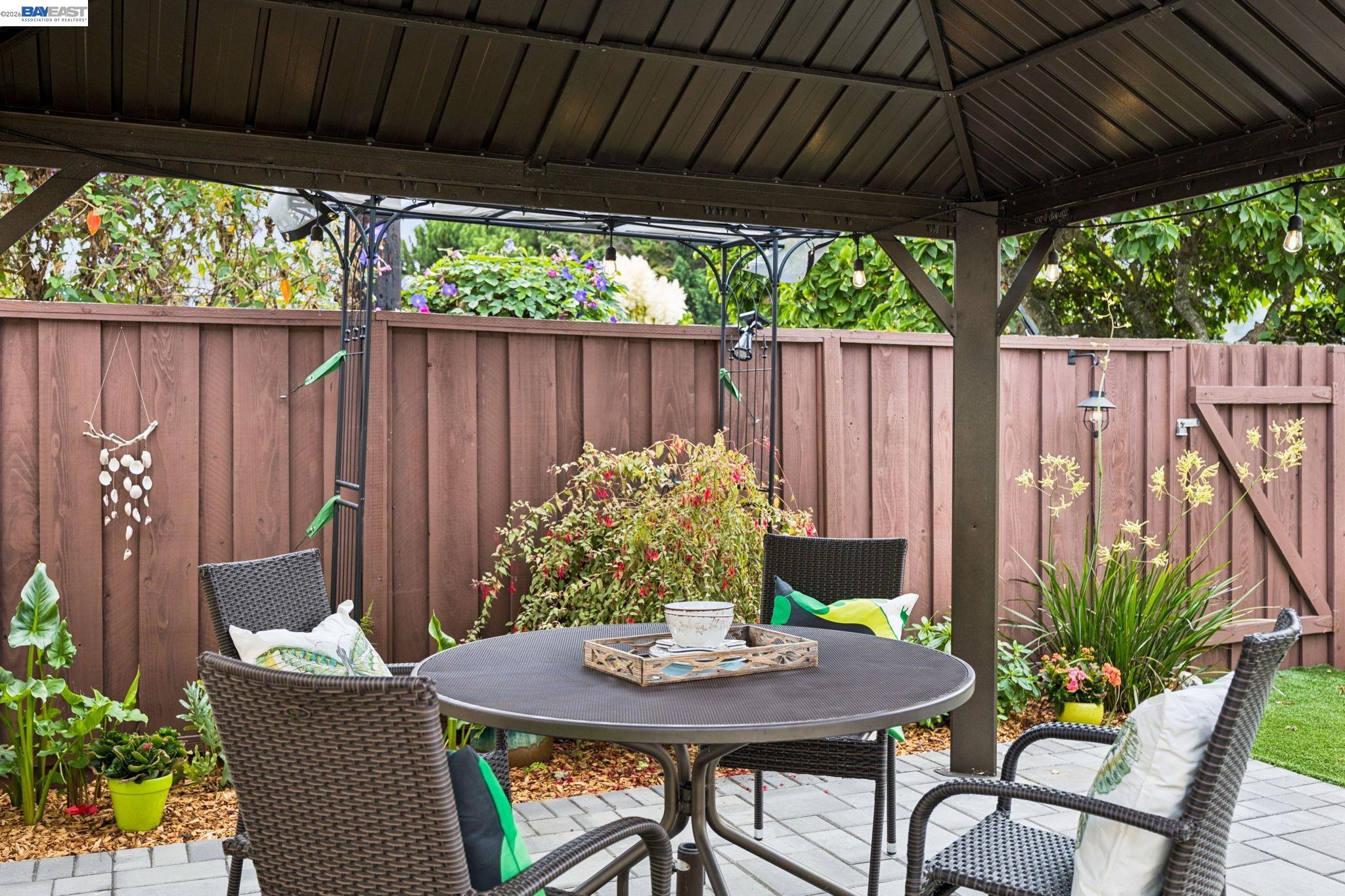 Covered backyard patio with wicker seating, a round table, flowering plants, and a tall wooden fence.