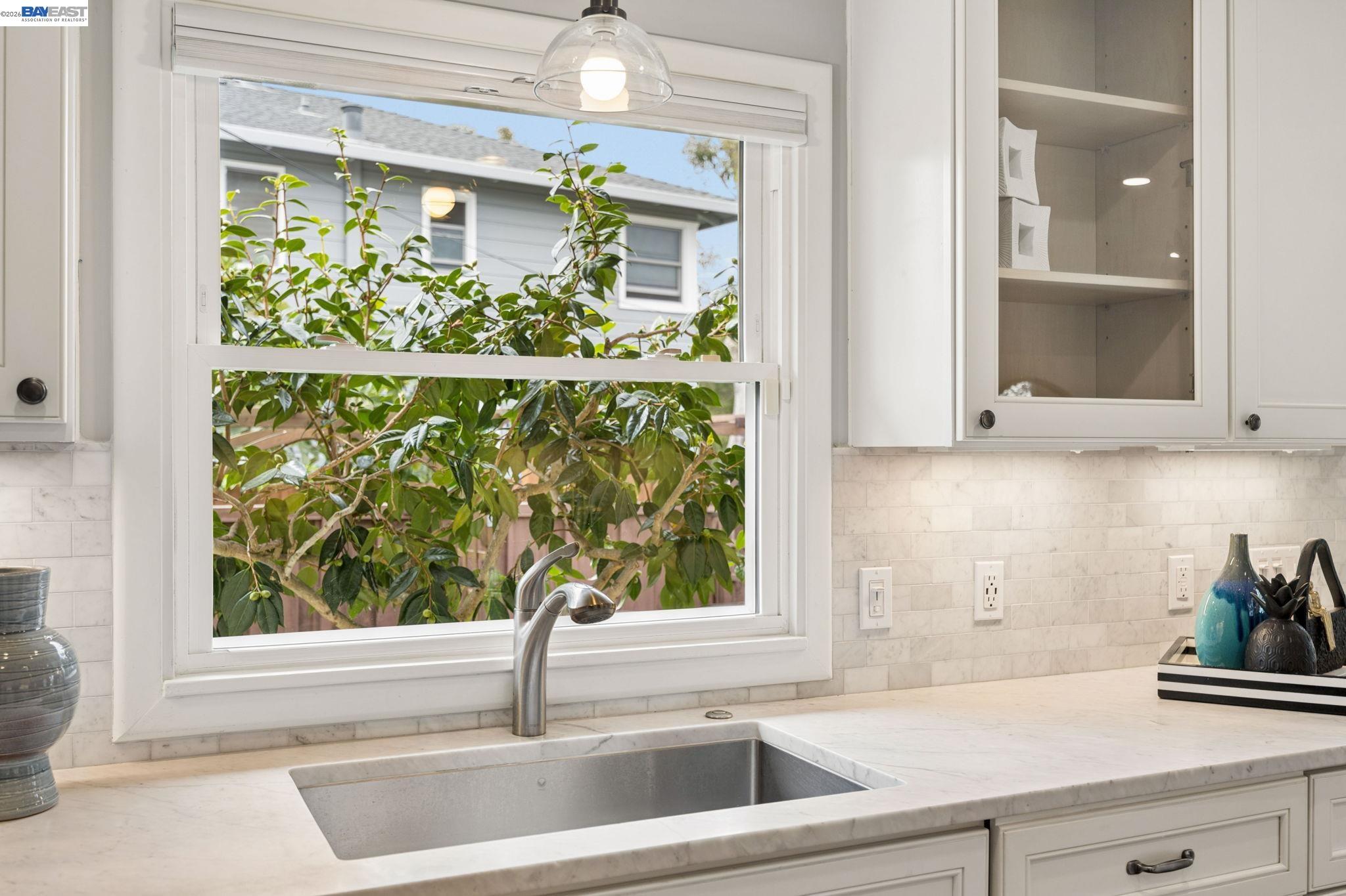 Bright kitchen sink beneath a large window, with leafy greenery outside and white marble counters.