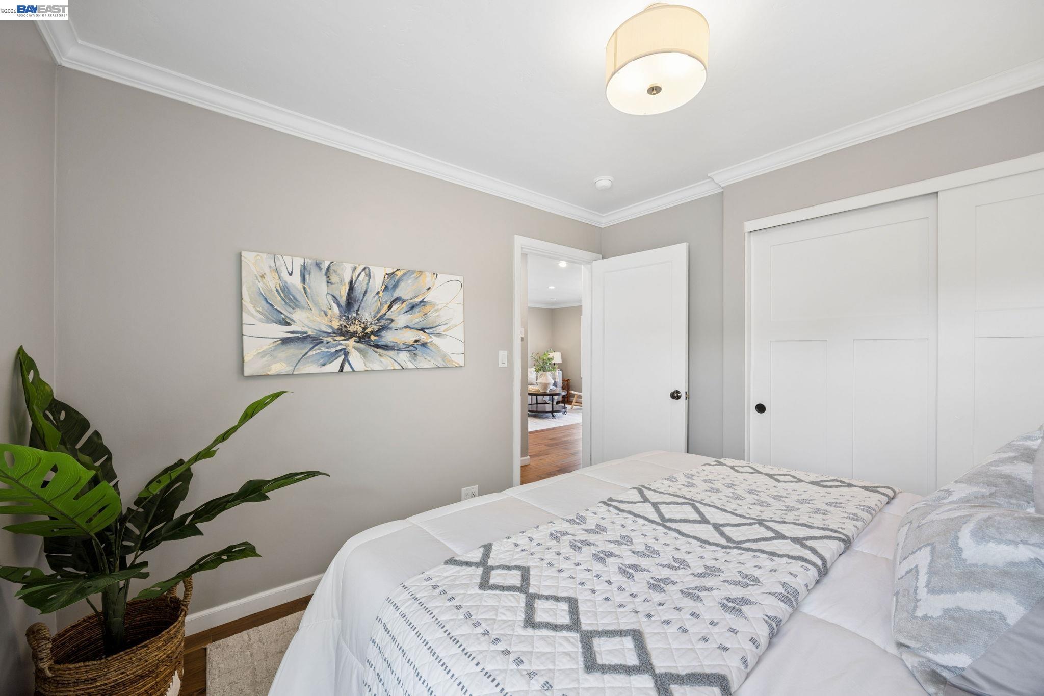 Bedroom with a white bed, gray geometric quilt, potted plant, and floral wall art.