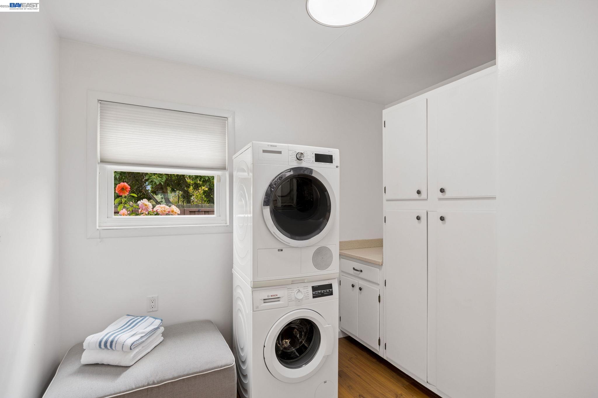 Laundry room with stacked washer and dryer, white cabinetry, and a flower-framed window.