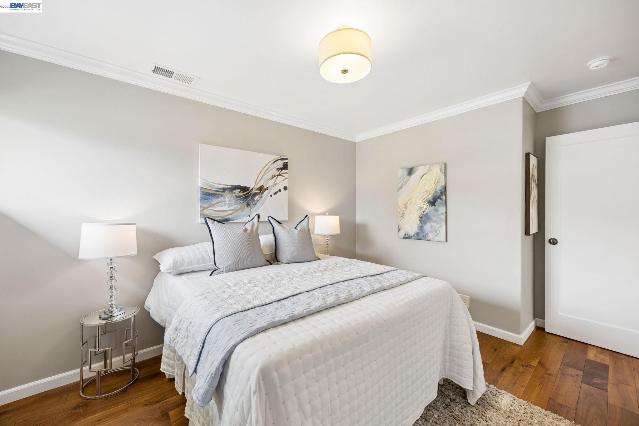 Staged bedroom with a white quilted bed, gray pillows, abstract art, and warm hardwood floors.