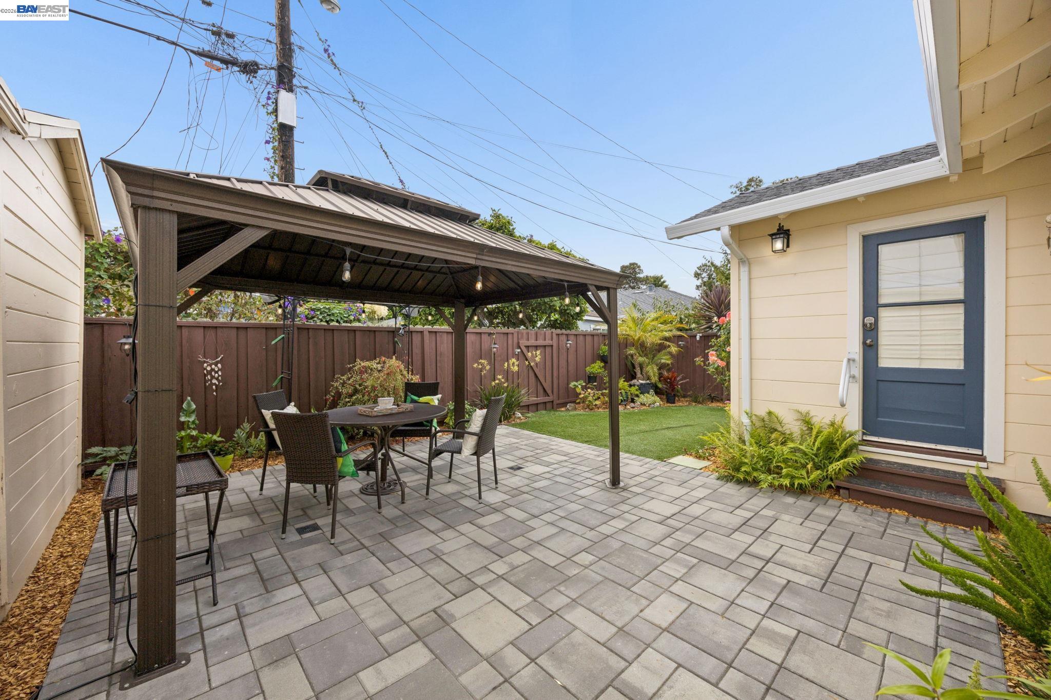 Paved backyard patio with dining set under a pergola beside a yellow house and small lawn.