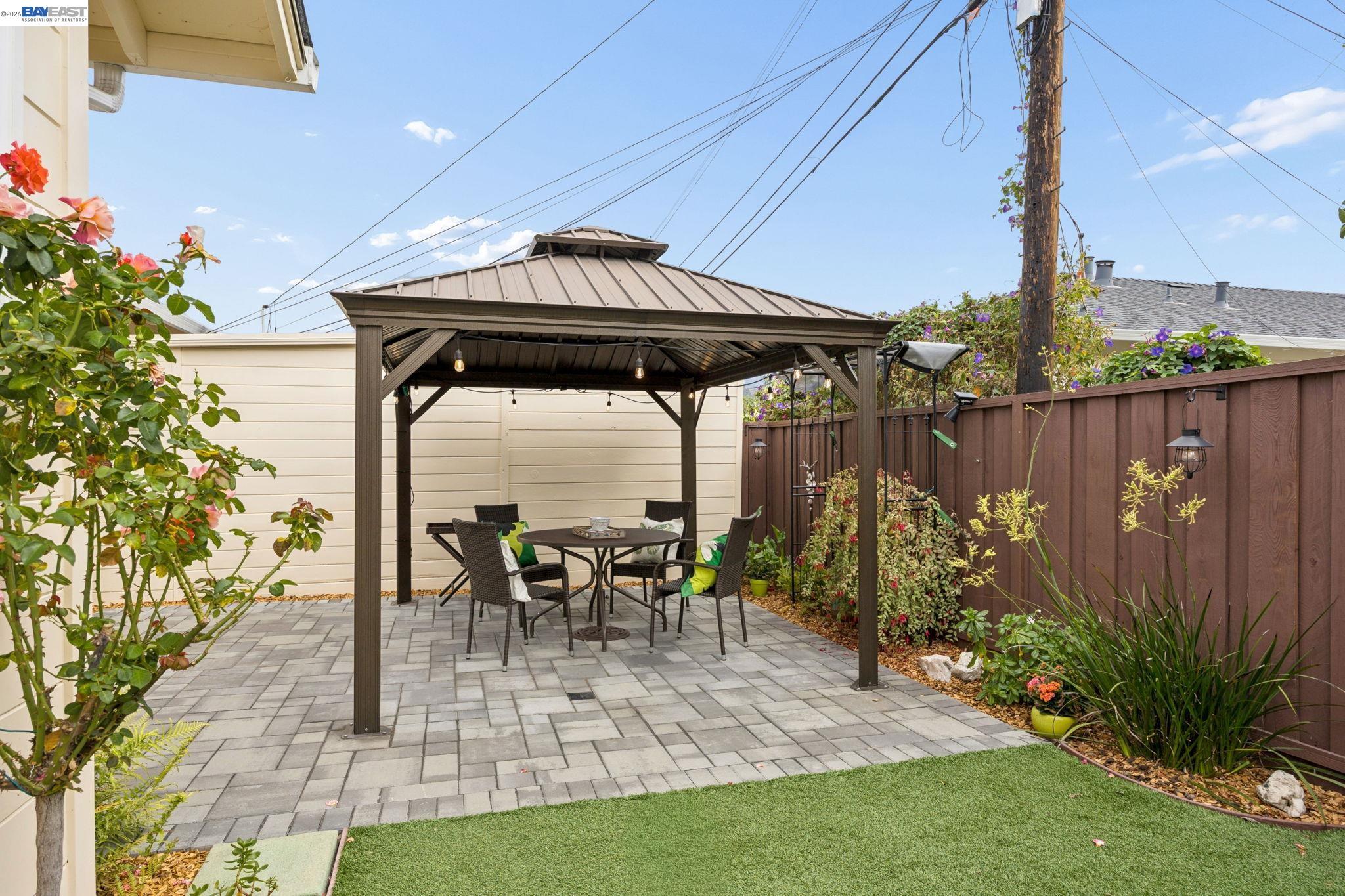 Backyard patio with a covered dining pavilion, paver flooring, artificial grass, and flowering garden beds.