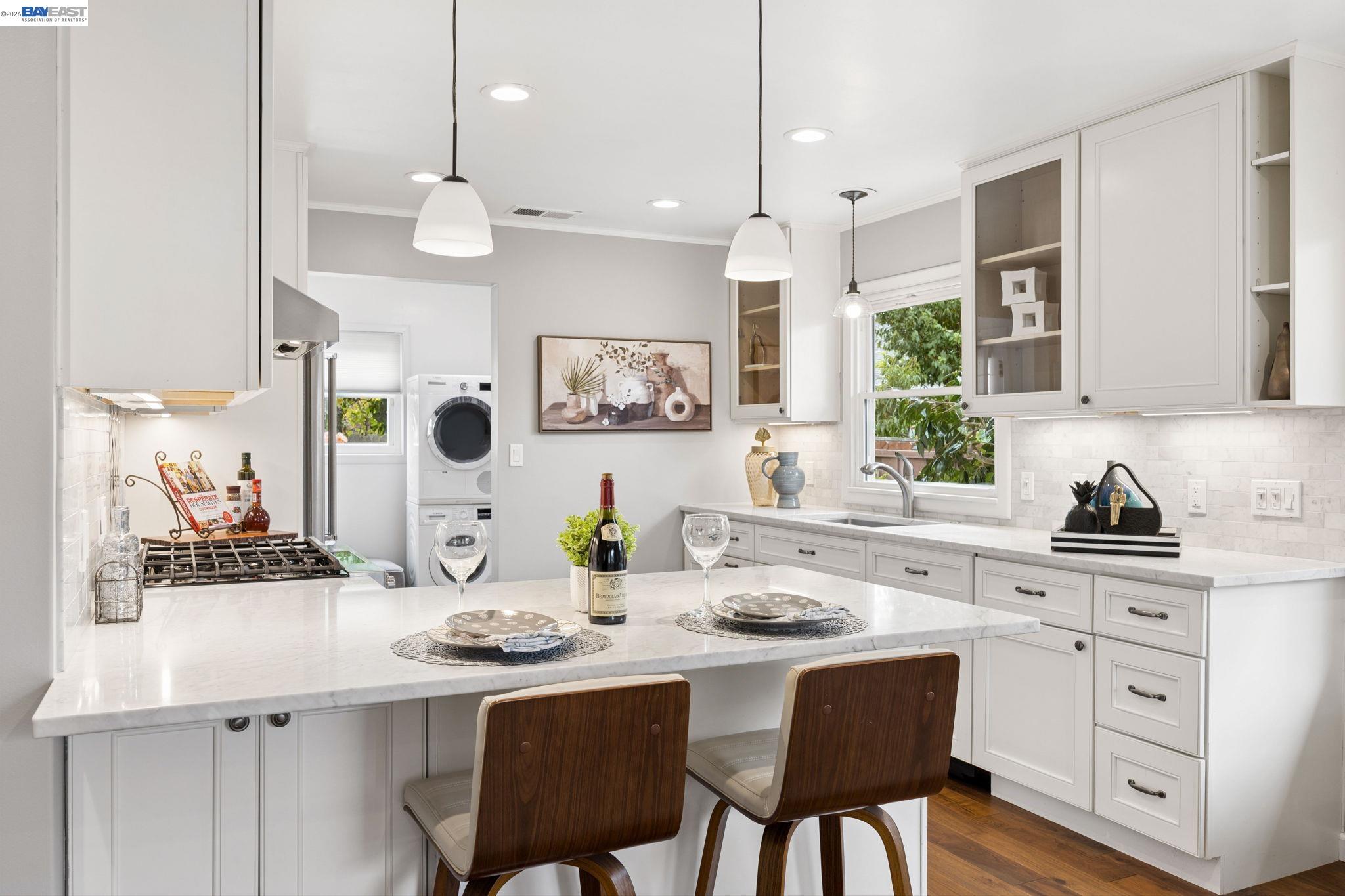Bright white kitchen with a large stone island, pendant lights, and stacked washer and dryer.