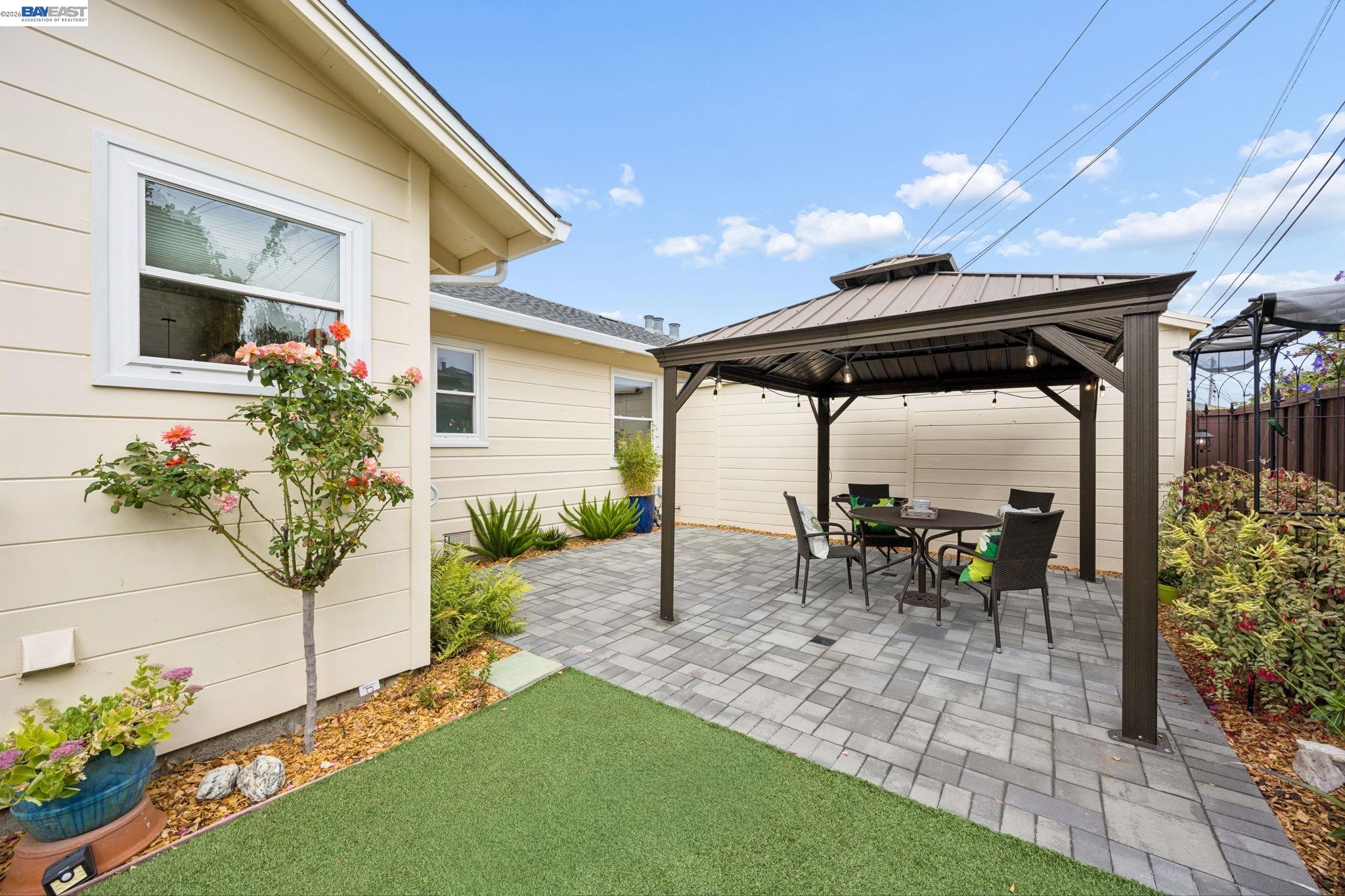 Backyard patio with a metal-roof gazebo, dining set, paver flooring, and flowering plants.