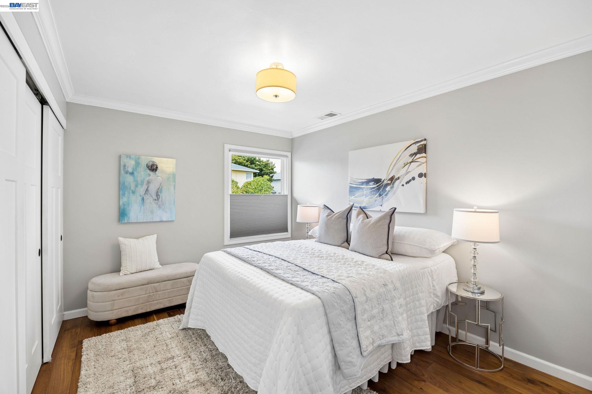 Staged bedroom with a white quilted bed, gray walls, hardwood floors, and a window.