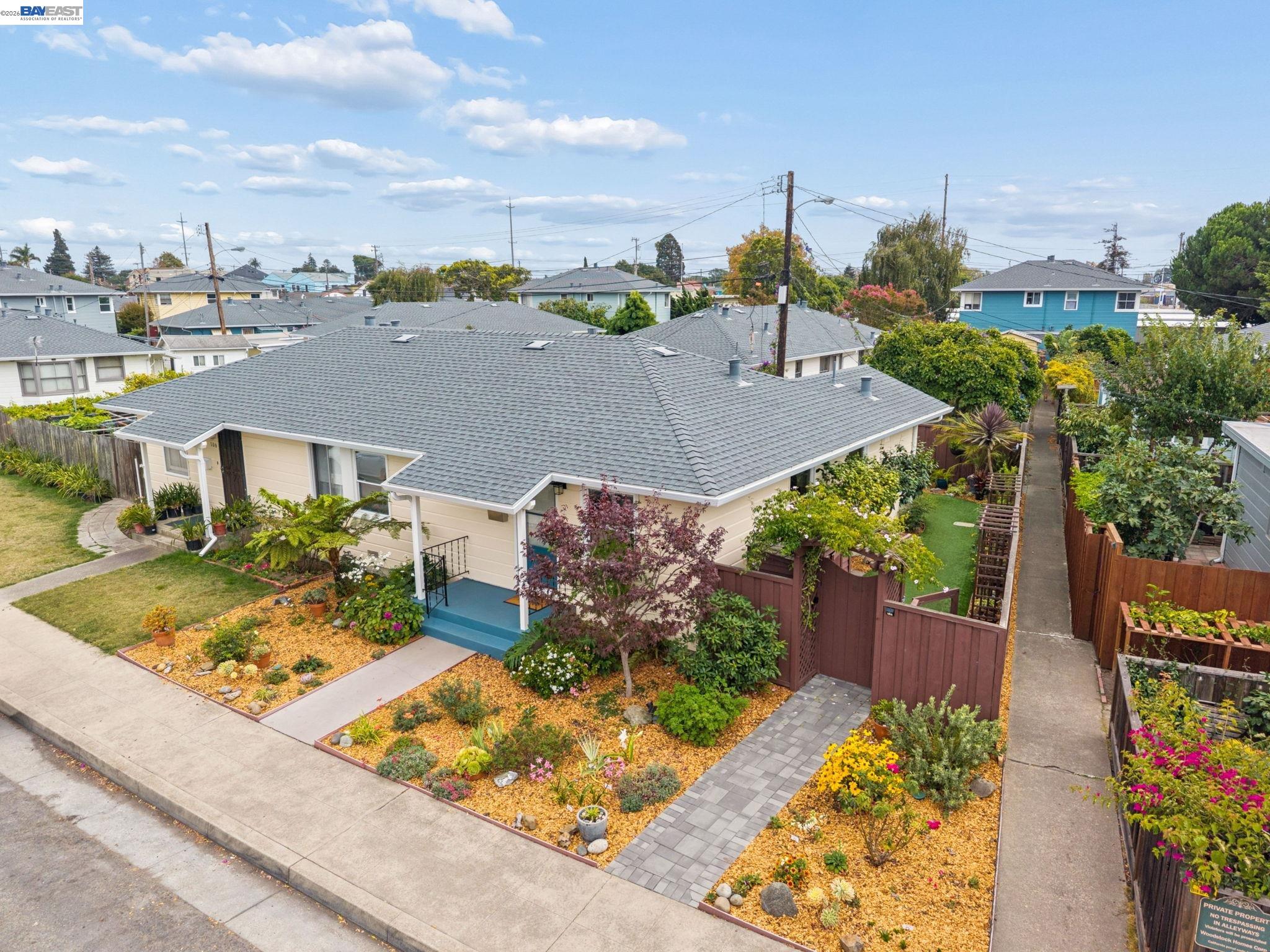 Single-story beige home with a broad gray roof, colorful landscaped yard, and paved walkway, viewed from above.