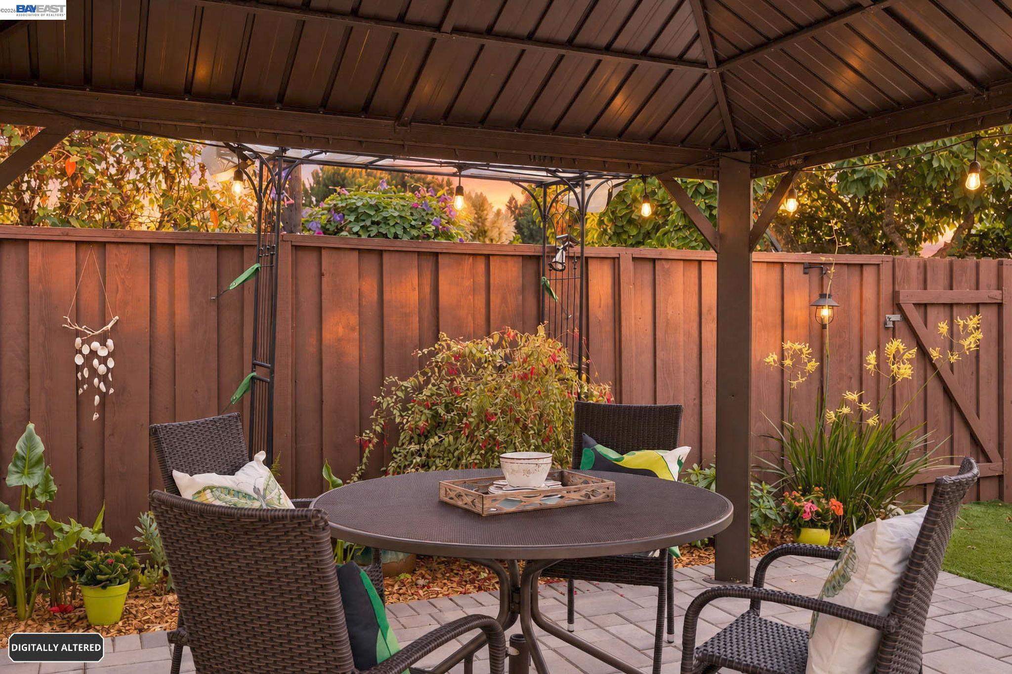 Covered backyard patio with wicker seating, string lights, and flowering plants.