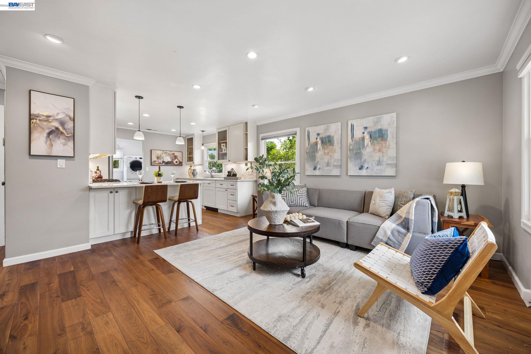 Open-plan living room and kitchen with a gray sectional, hardwood floors, and a breakfast counter with two stools.
