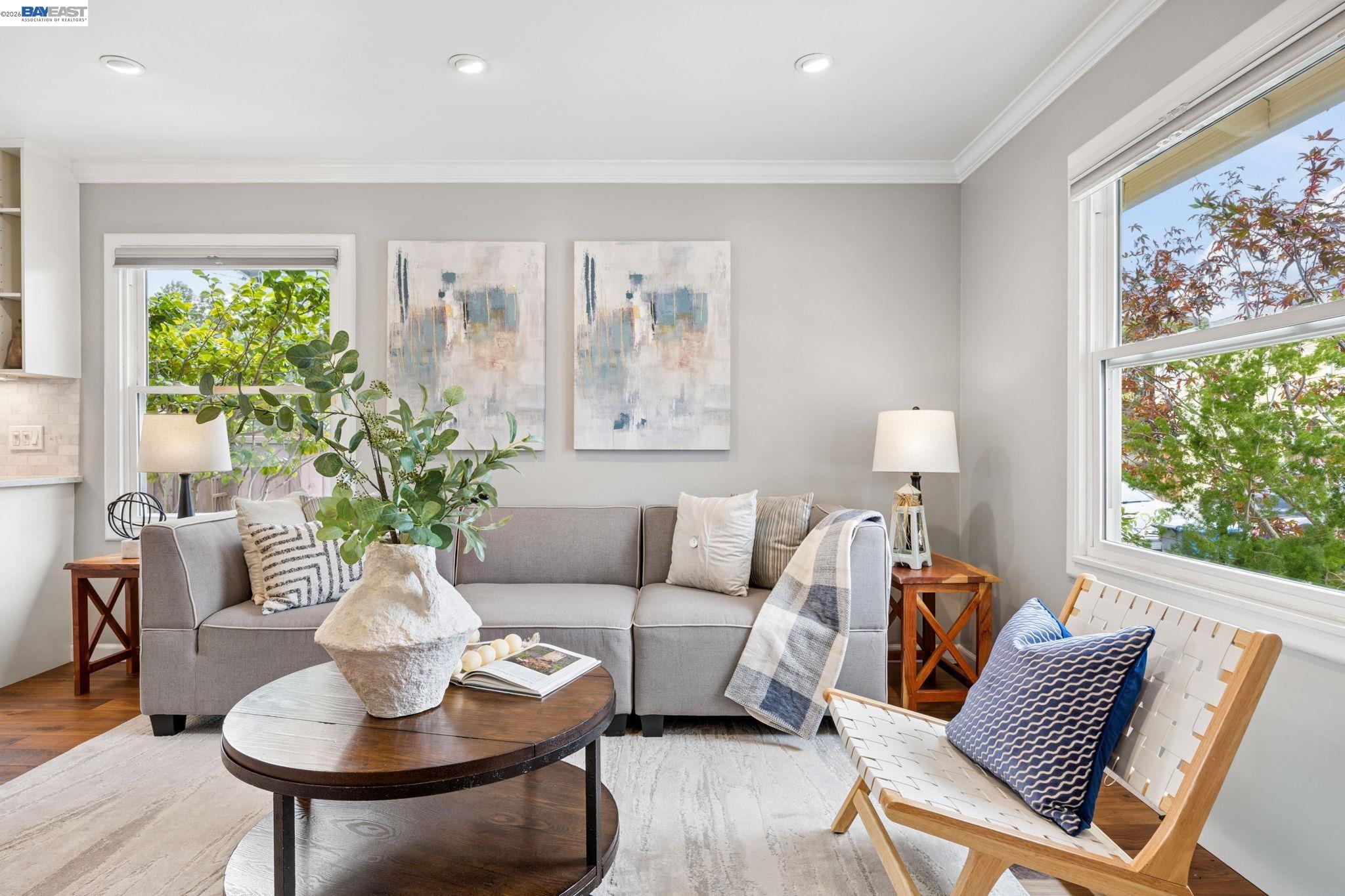 Staged living room with a gray sofa, abstract art, and large windows overlooking leafy trees.