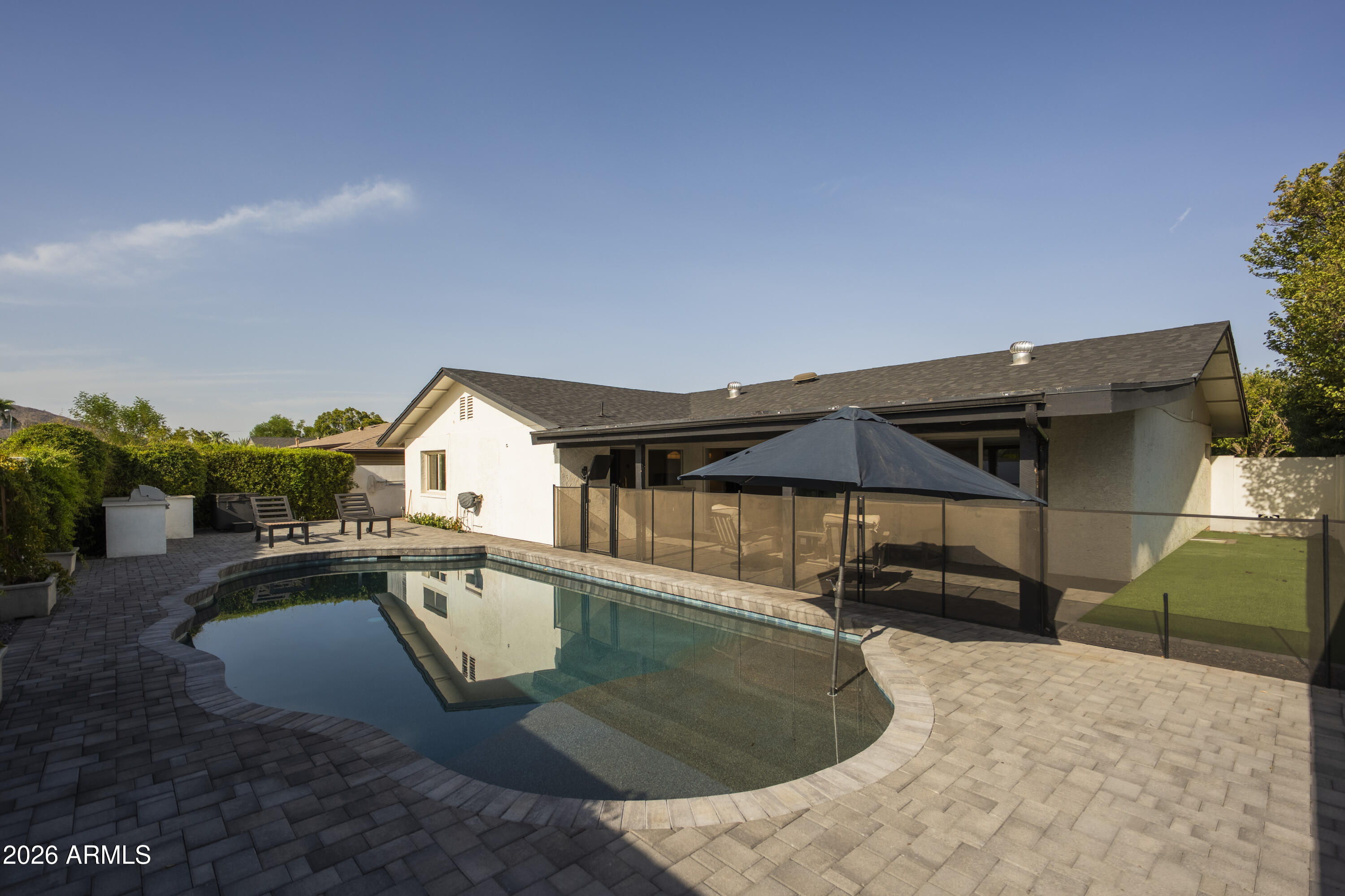 Home’s backyard with a curved swimming pool, paved patio, and shaded seating area.
