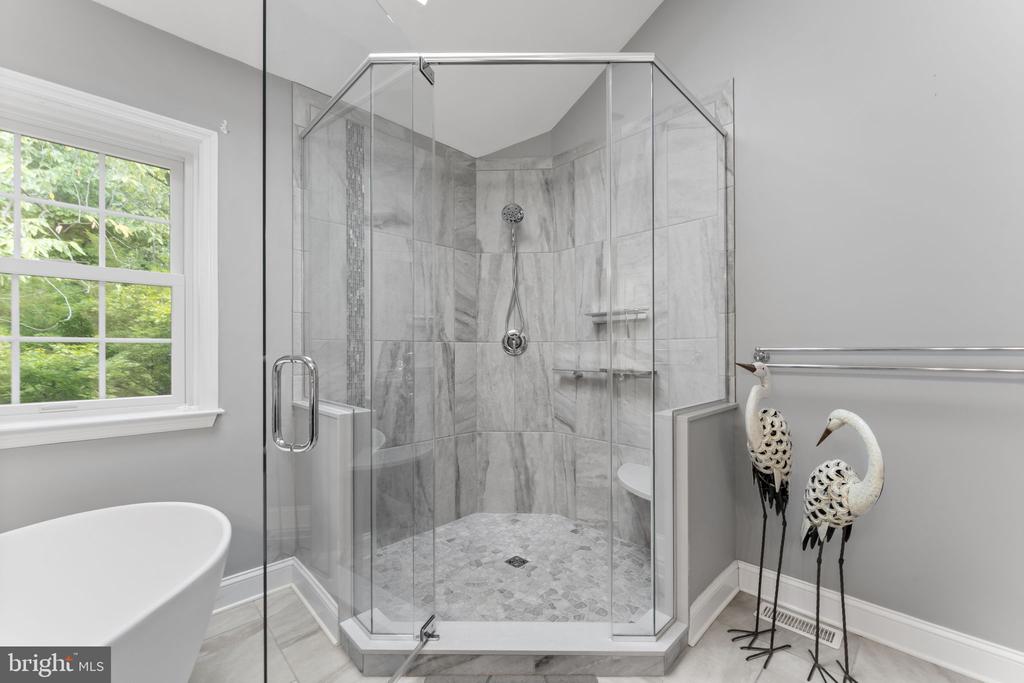 Bathroom with a marble-look glass shower, freestanding tub, and window overlooking greenery.