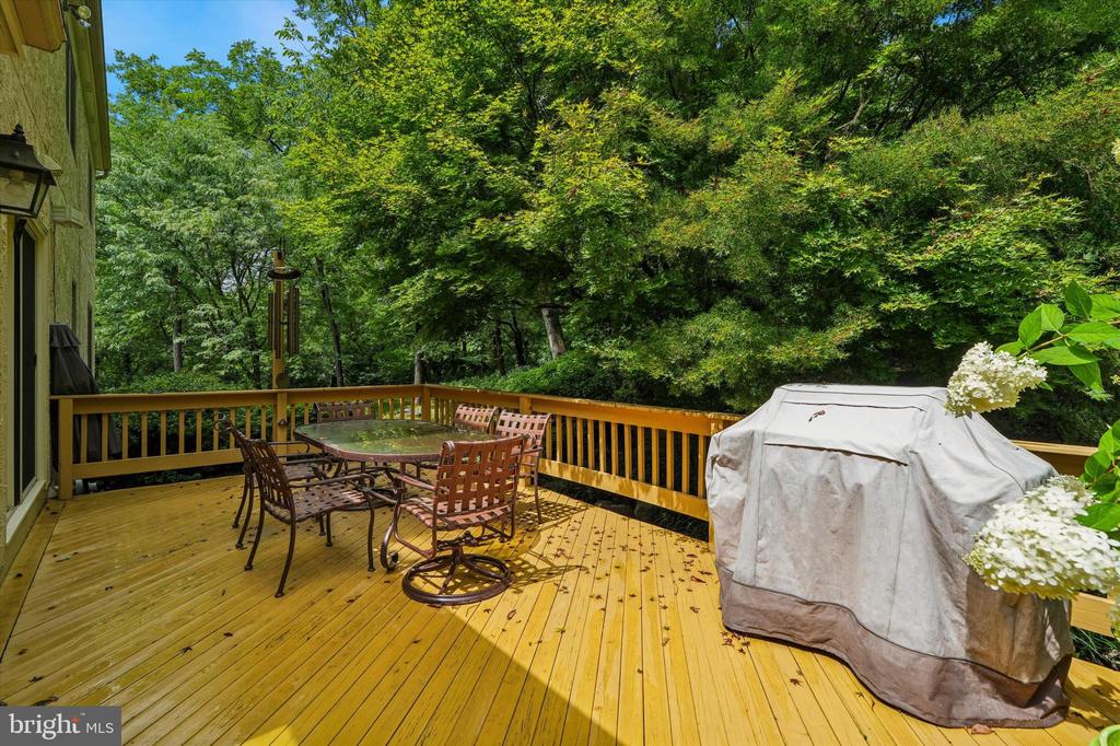 Wooden deck with a patio dining set and covered grill, surrounded by leafy trees.