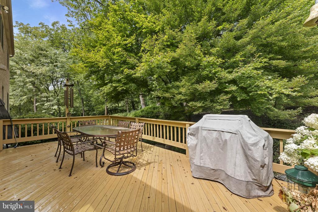 Wooden deck with patio dining set and covered grill, surrounded by leafy trees.