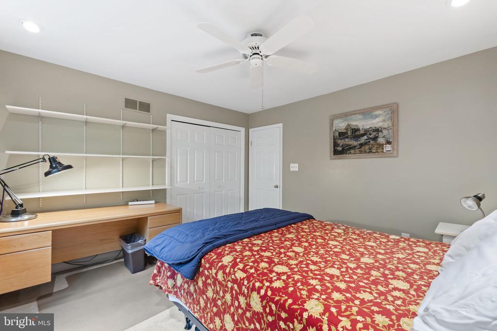 Bedroom with a red floral bedspread, blue blanket, and wall shelving above a wooden desk.