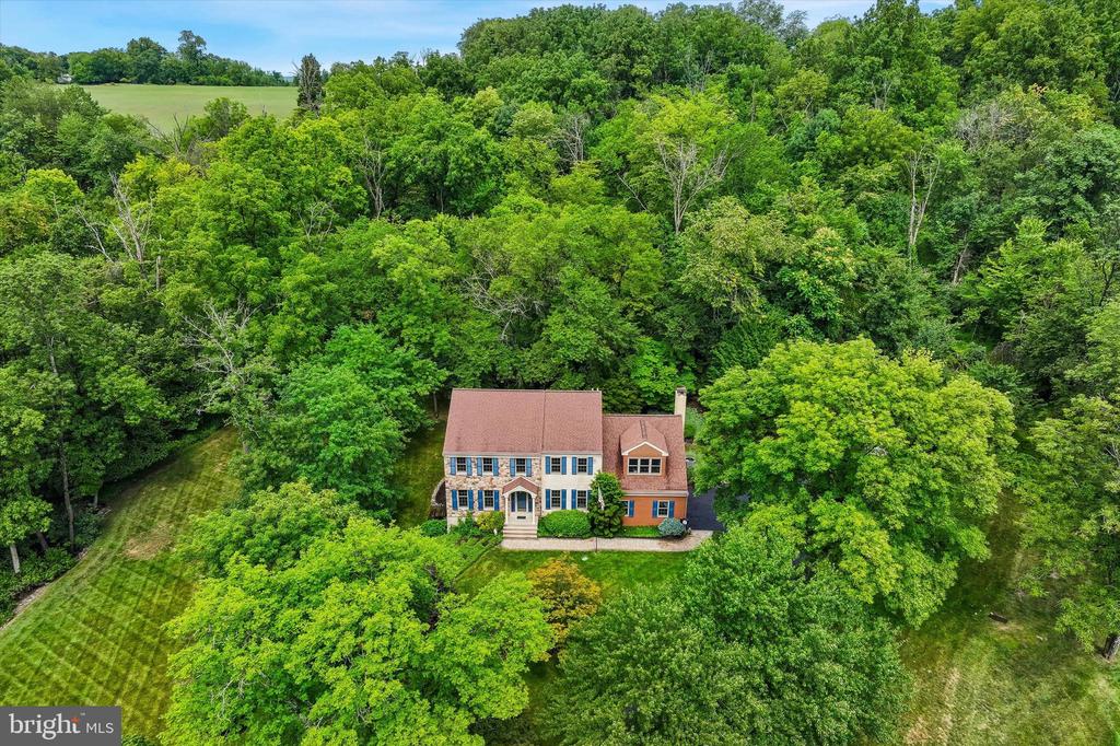 Aerial view of a two-story house nestled among trees, with a broad lawn and open field beyond.