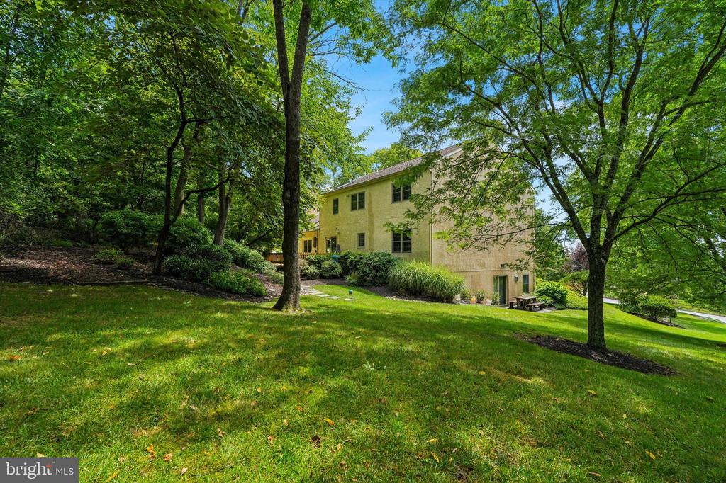 Two-story beige house behind a broad lawn, framed by mature leafy trees.