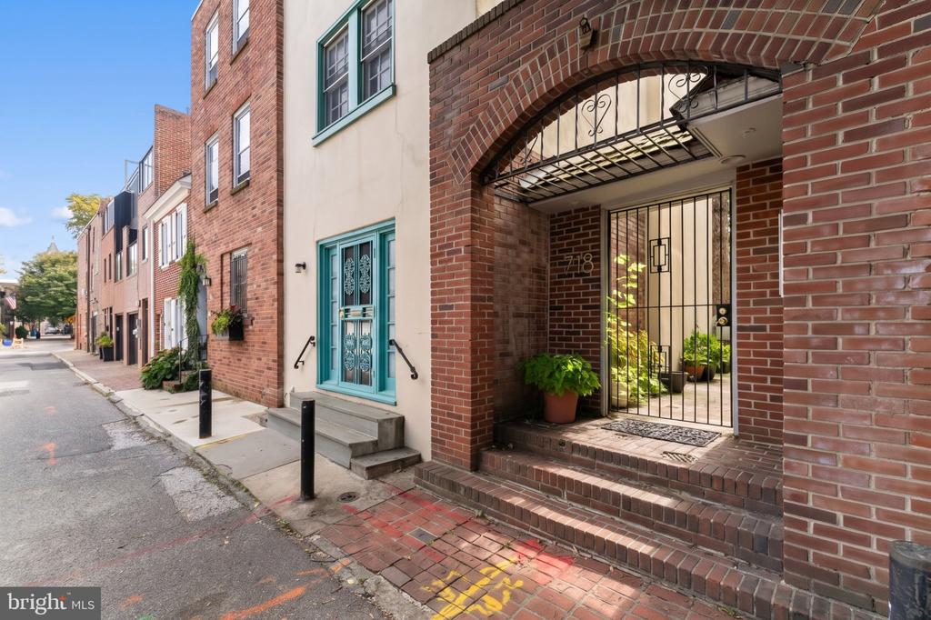 Brick rowhouse entrance with an arched canopy, iron gate, potted plants, and turquoise neighboring doorway.