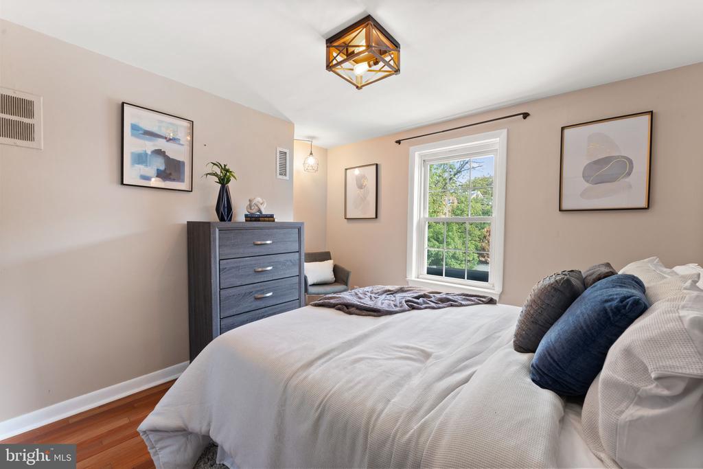 Furnished bedroom with a white bed, blue pillows, gray dresser, and window overlooking trees.