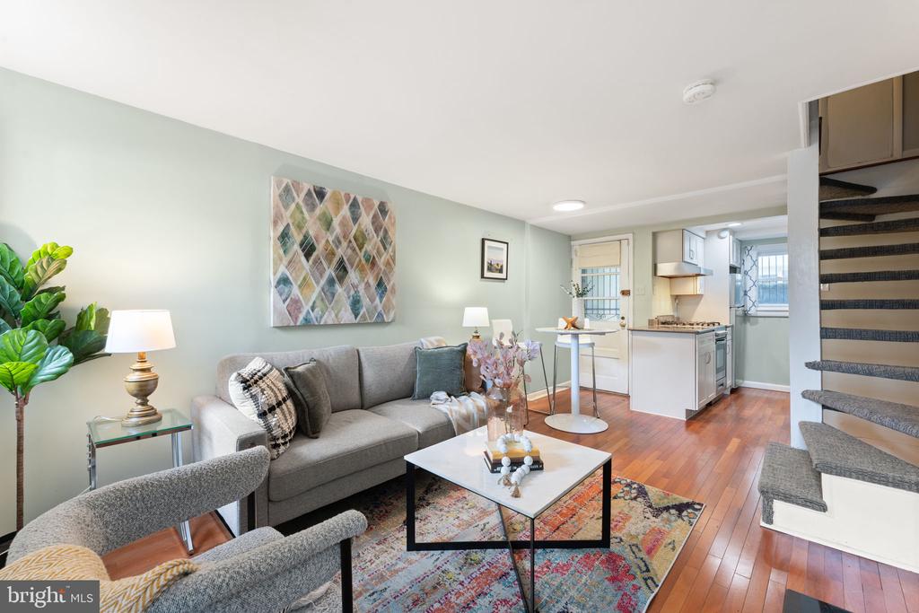 Staged living room with gray sofa, colorful rug, open kitchen, and staircase.