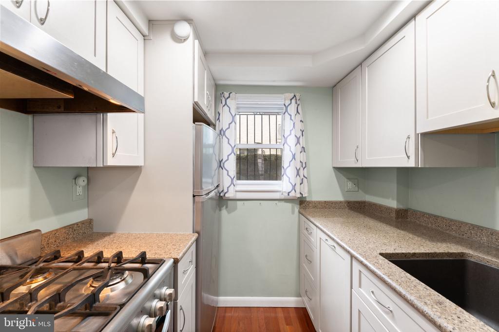 Galley kitchen with white cabinets, stone countertops, stainless-steel appliances, and a barred window with patterned curtains.