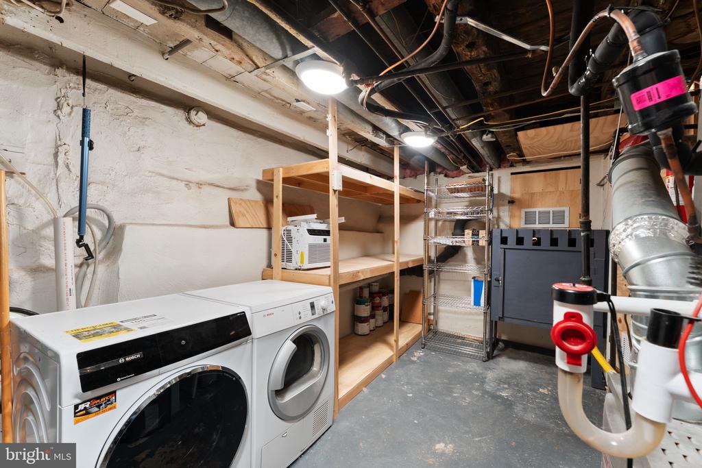 Basement laundry room with Bosch washer and dryer, open wooden shelves, and exposed utility pipes.