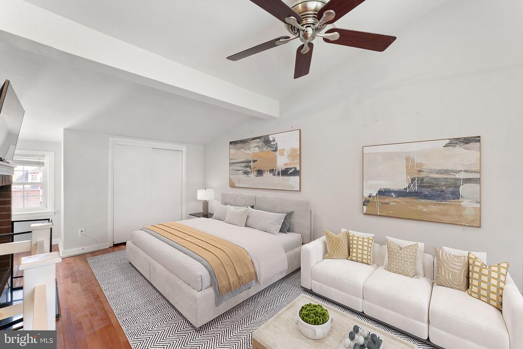 Staged bedroom with gray upholstered bed, white sectional sofa, patterned rug, and ceiling fan.