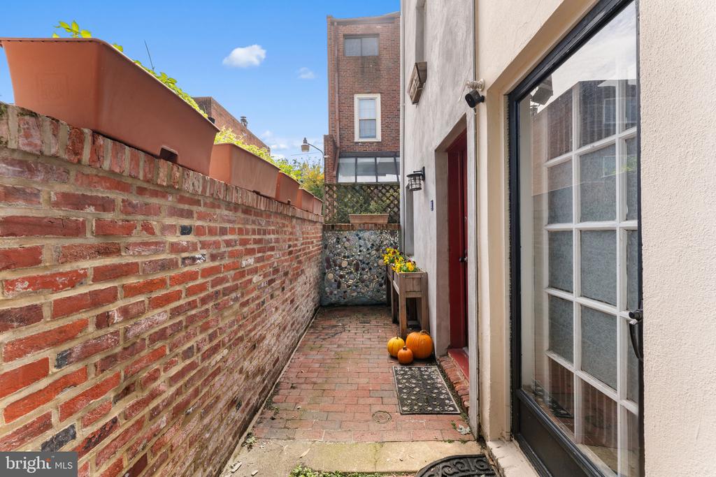 Brick-paved side patio with a long brick wall, glass door, flower table, and pumpkins by the entrance.