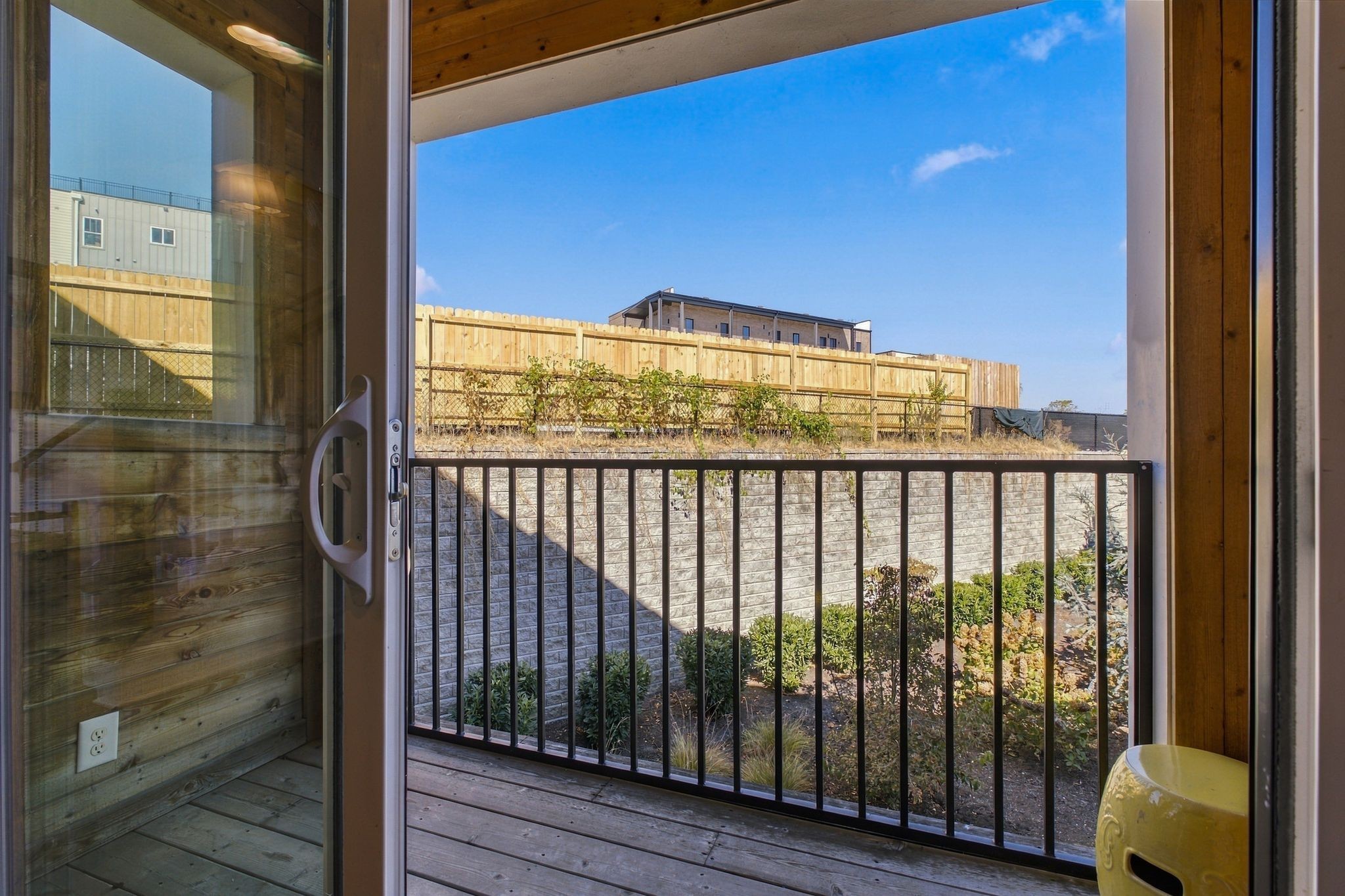 Wood-deck balcony with black railing and yellow ceramic stool overlooking a fenced yard.