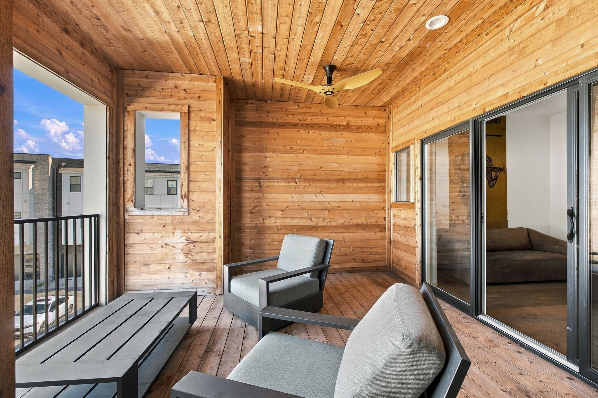 Covered wooden balcony with gray seating, a ceiling fan, and sliding doors to the living room.