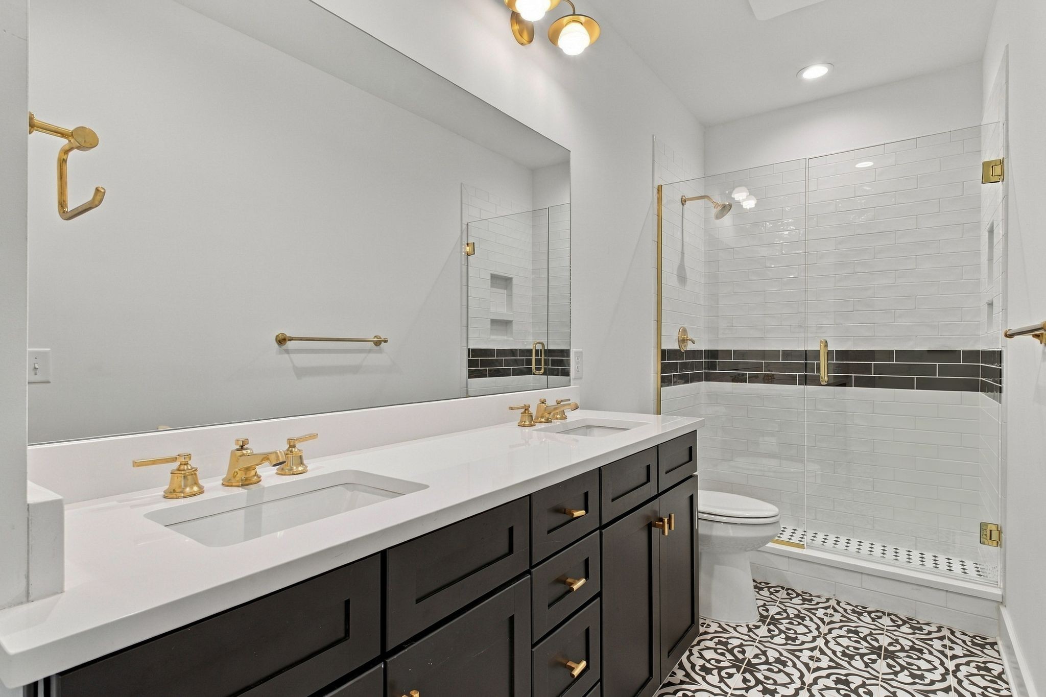 Bathroom with a dark double vanity, gold fixtures, glass shower, and patterned floor tiles.
