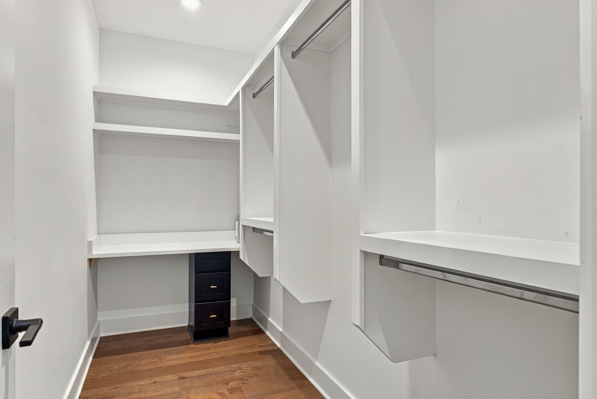 Walk-in closet with white built-in shelves, hanging rods, dark drawers, and wood flooring.
