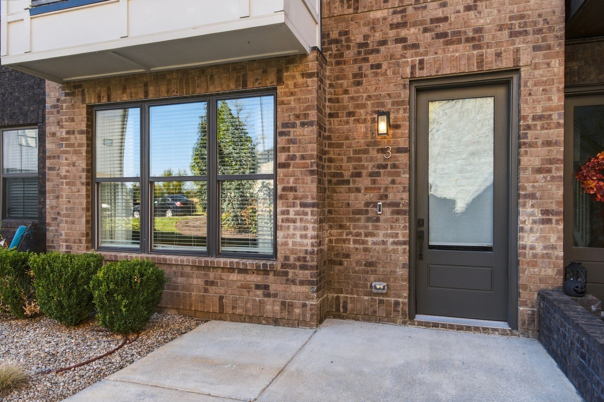 Brick townhome entrance with a dark door marked 3, large windows, and trimmed shrubs.