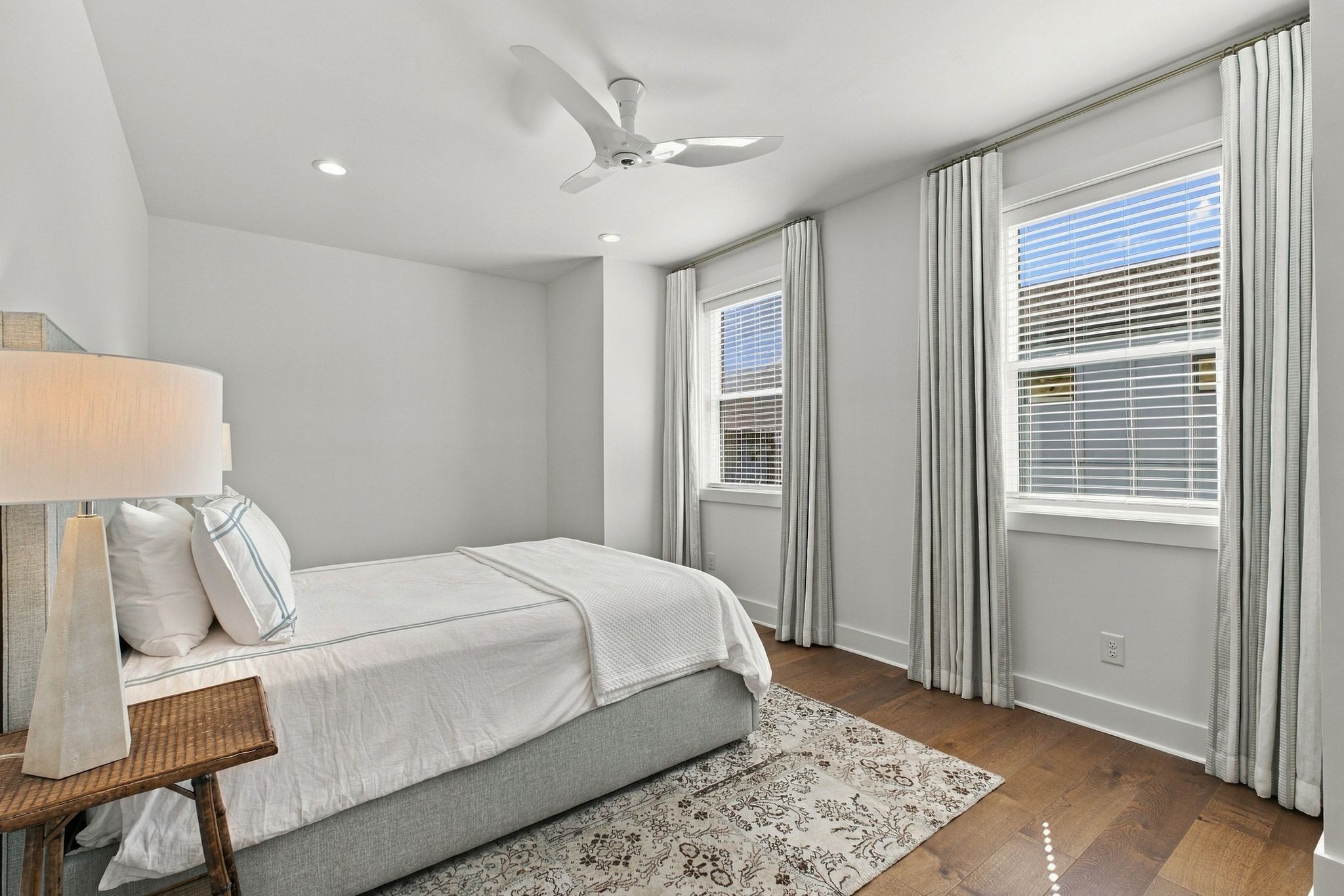 Bright bedroom with a neatly made bed, two curtained windows, a ceiling fan, and hardwood floors.