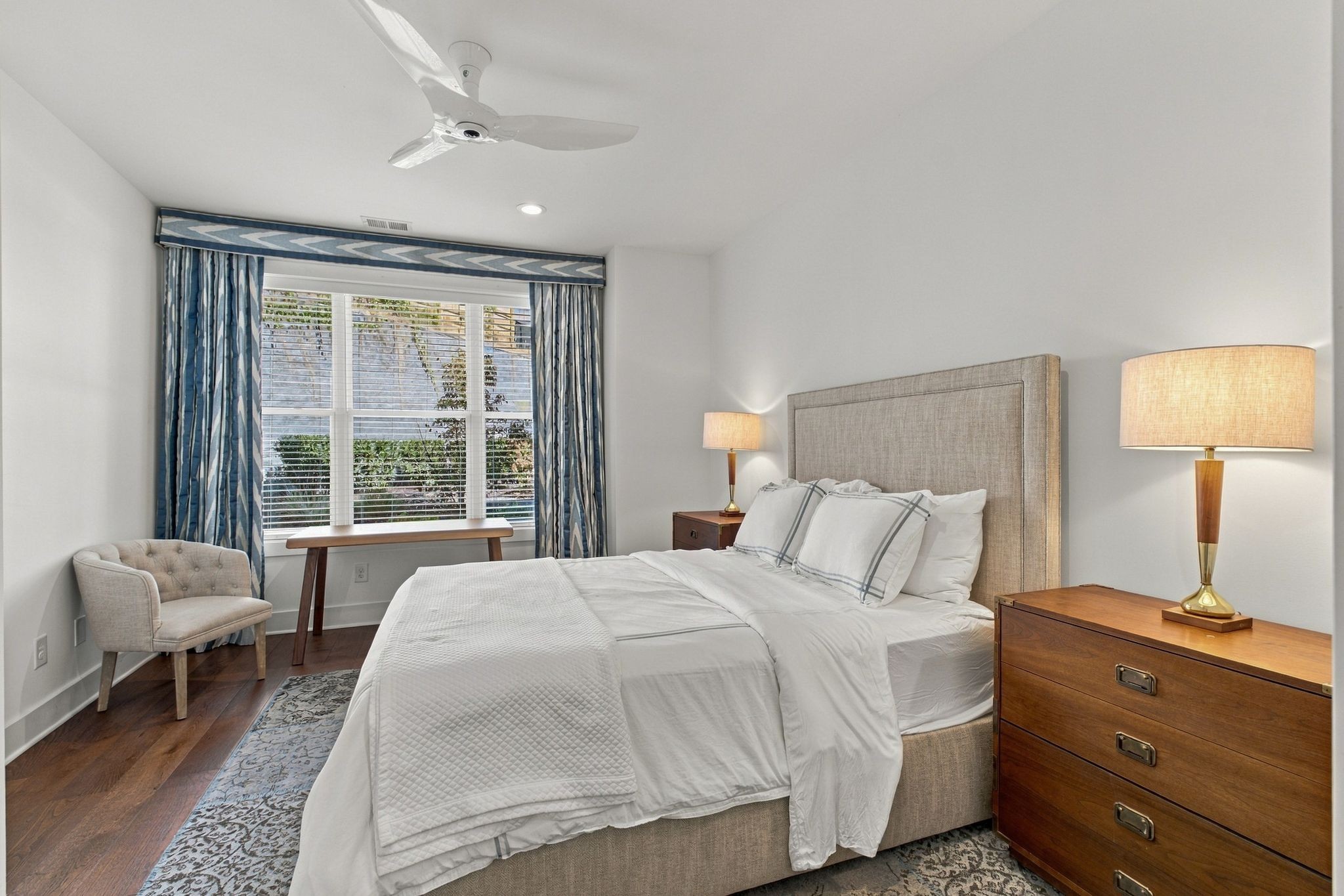 Staged bedroom with a white bed, upholstered headboard, and blue curtains beside a window.