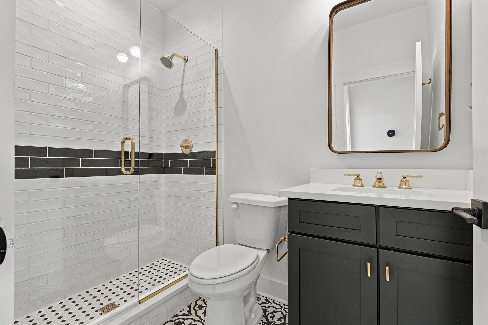 Bathroom with a glass shower, black accent tile, patterned floor, and dark vanity with brass fixtures.