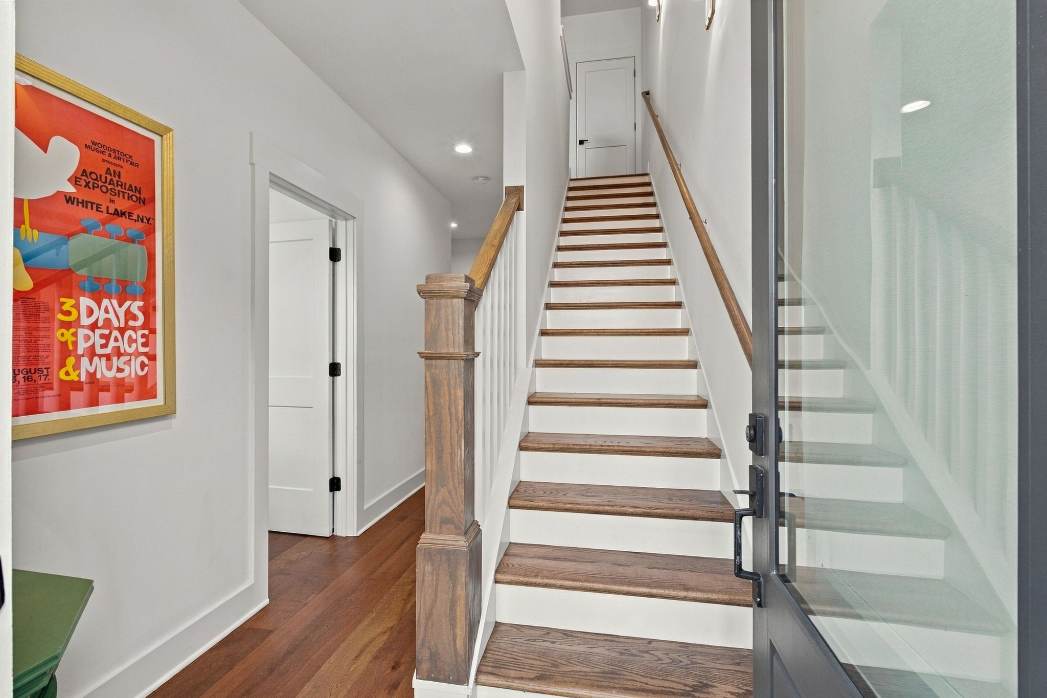 Entry staircase with dark wood treads, wood railings, glass-panel door, and framed Woodstock poster.