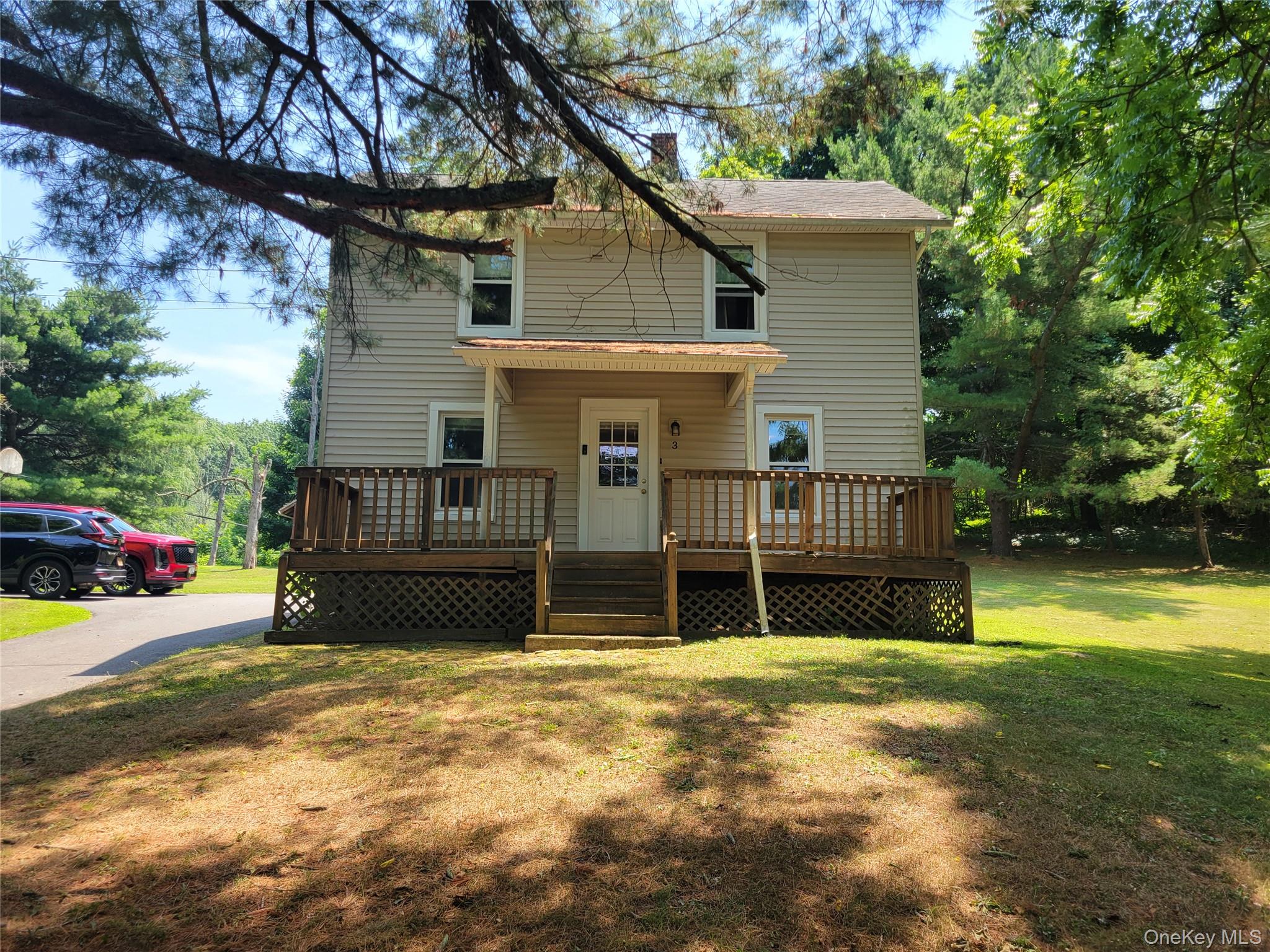 Two-story beige house with a raised wooden porch, surrounded by mature trees.