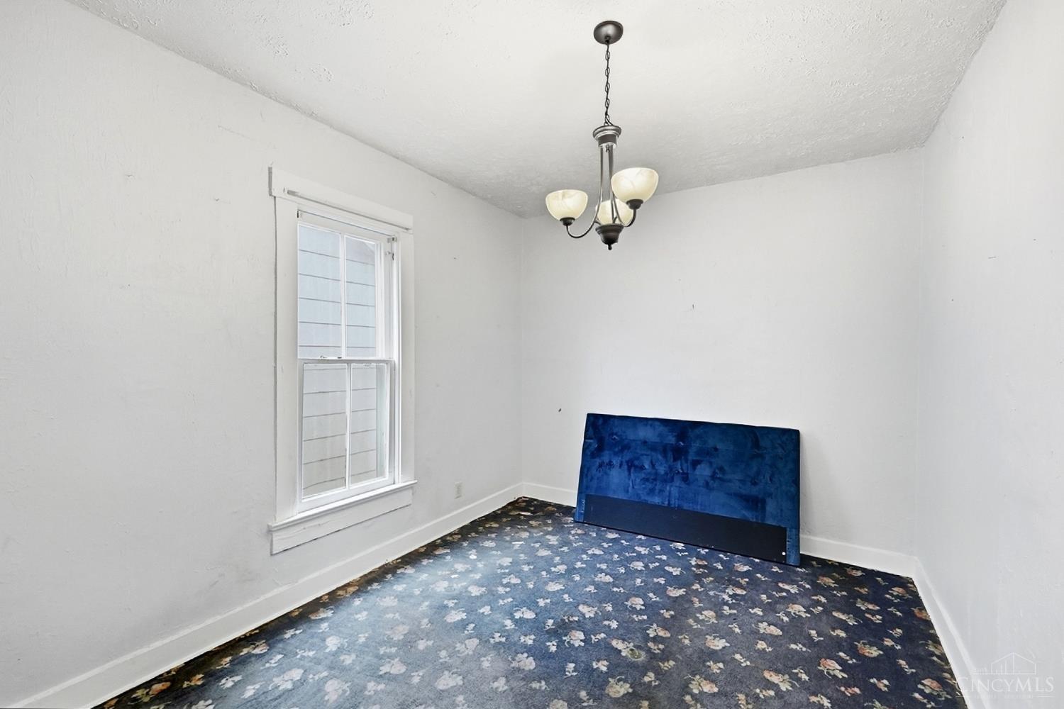 Mostly empty bedroom with a window, hanging chandelier, and blue floral carpet.
