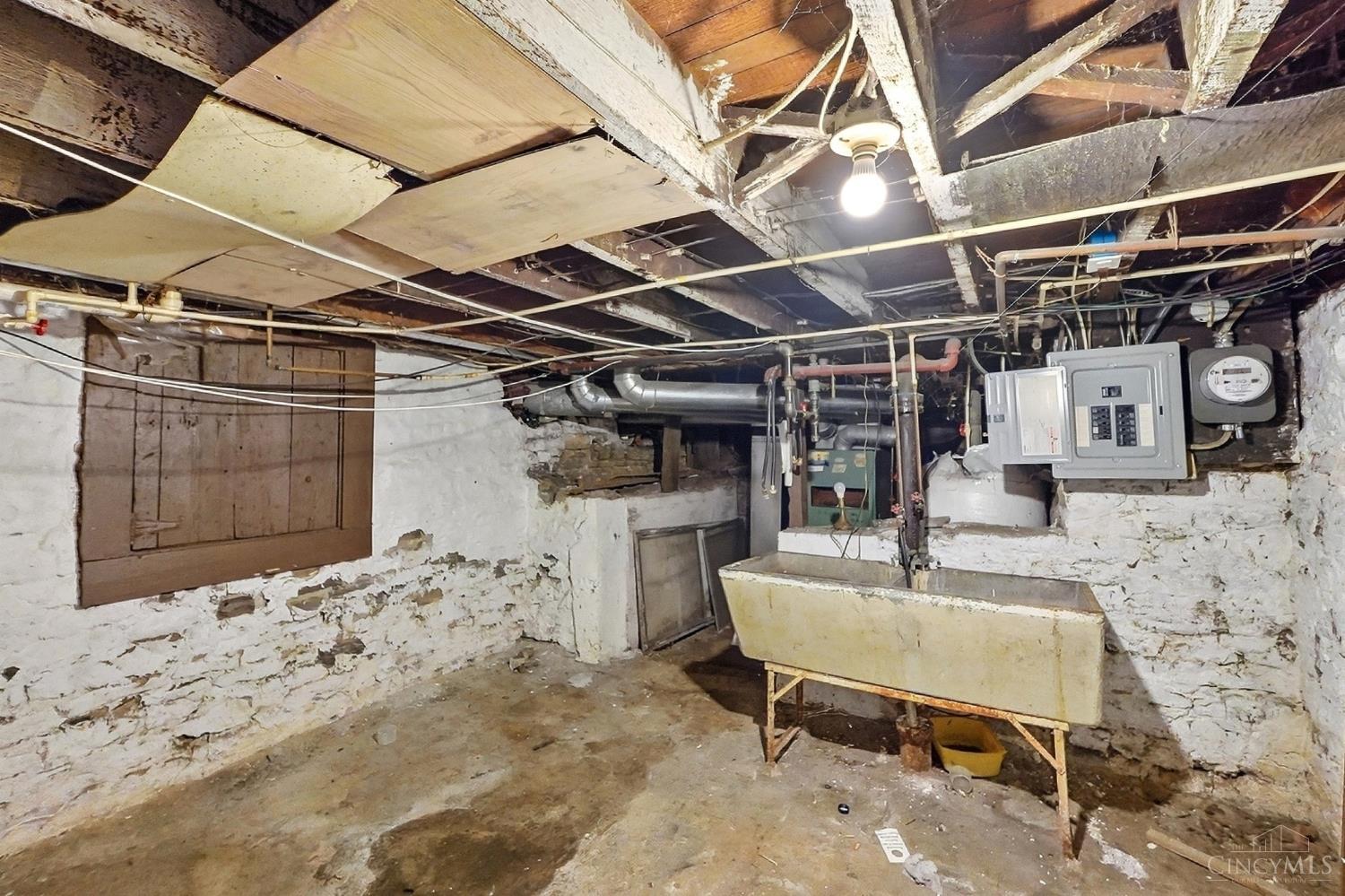 Unfinished basement with exposed joists, stone foundation walls, electrical panel, and utility sink.
