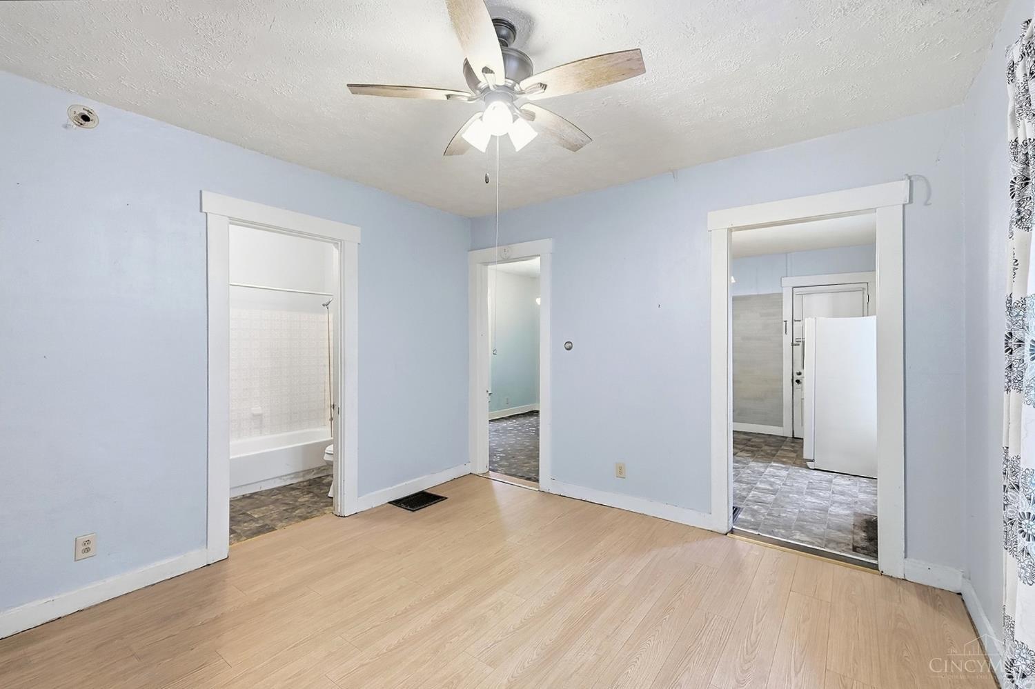 Empty pale blue room with light wood flooring, ceiling fan, and doorways to adjoining rooms.