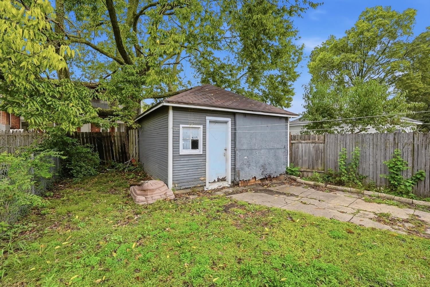 Gray backyard shed with a white door and window beside a concrete patio.
