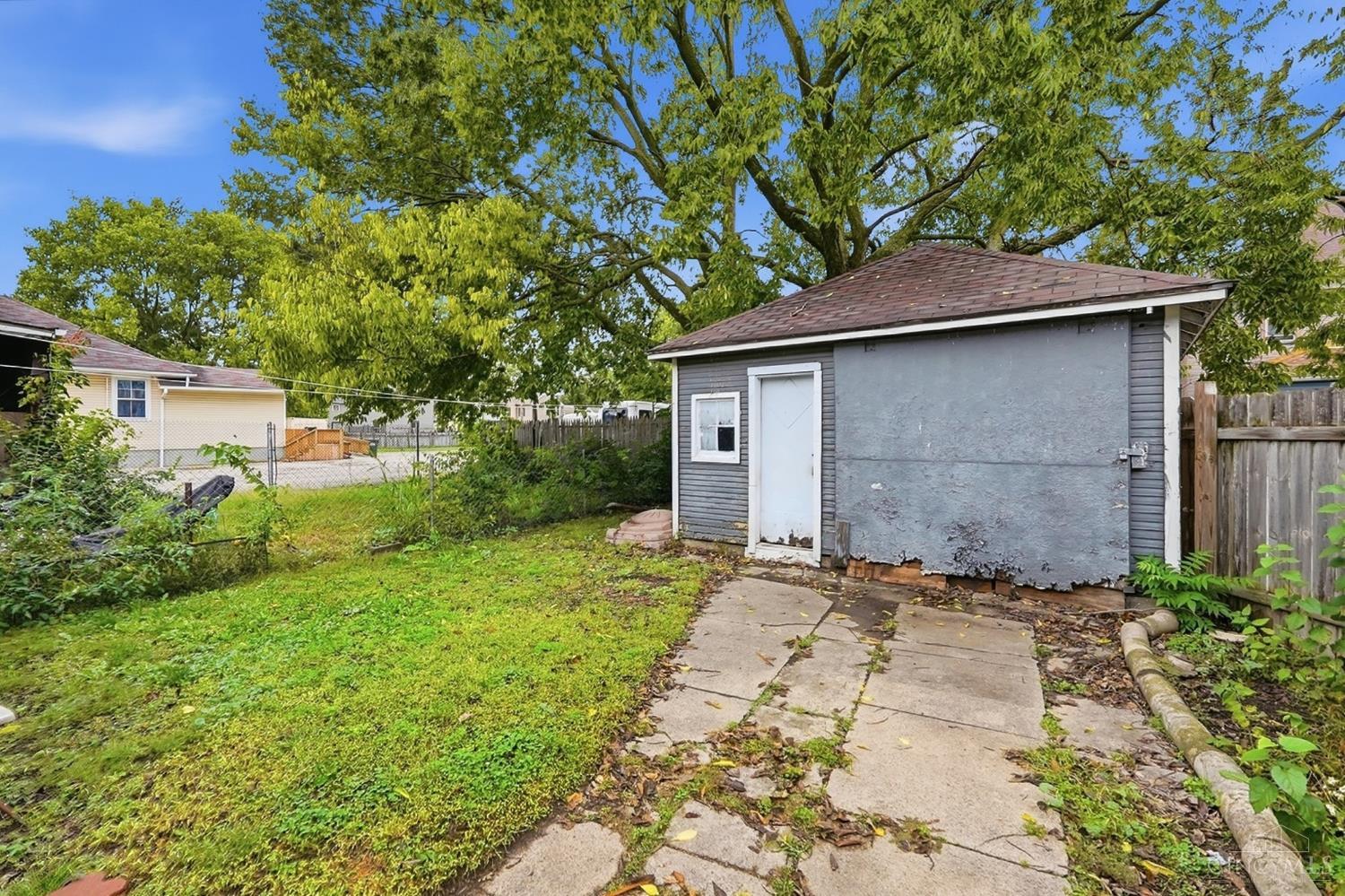 Backyard with a gray detached outbuilding, cracked concrete walkway, grassy lawn, and mature trees.