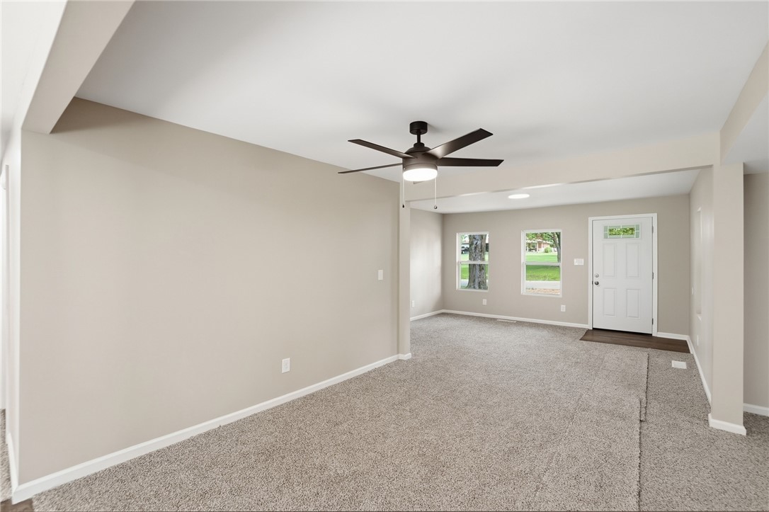 Empty living room with gray carpet, ceiling fan, and front door beside two windows.