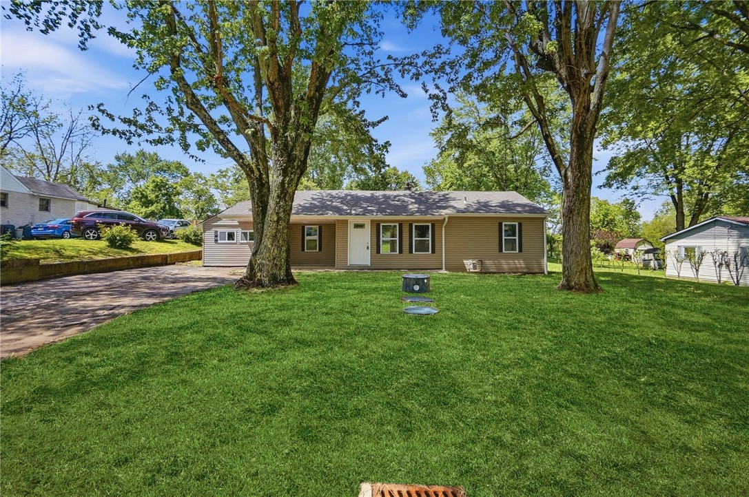 Single-story tan home with a white front door, broad lawn, mature trees, and driveway.