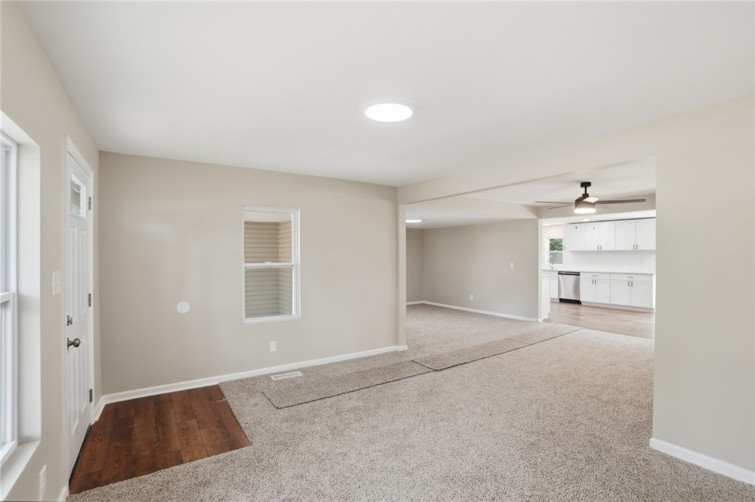 Vacant living area with beige carpet, a hardwood entry, and the white kitchen visible beyond.
