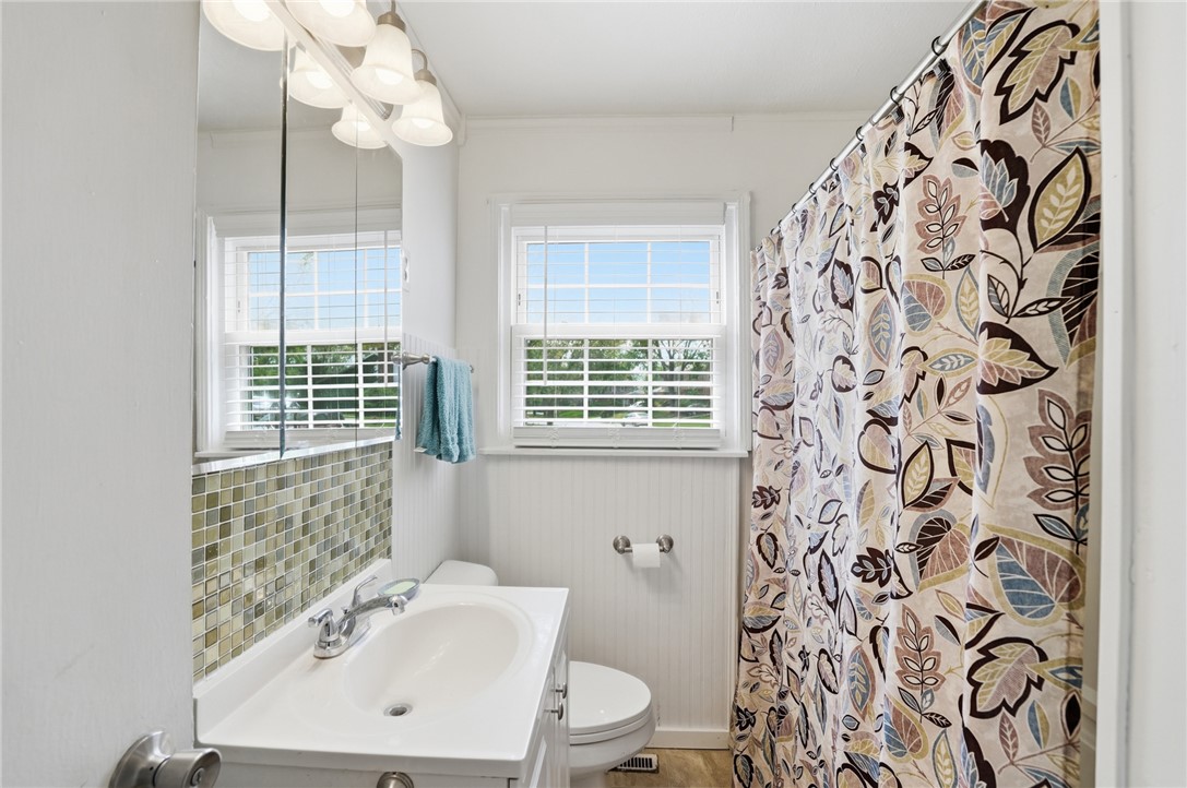 Bathroom with a white vanity, green mosaic backsplash, and patterned shower curtain beside two windows.